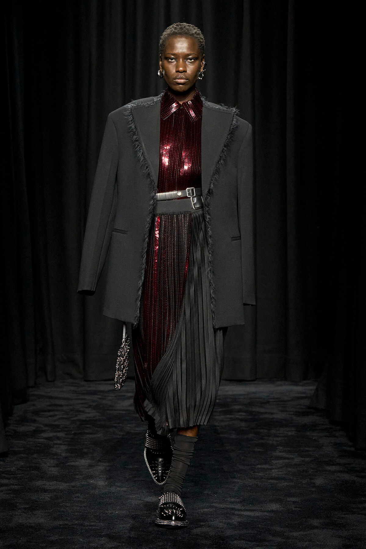 Fashion model walking the runway in a black fringed blazer over a burgundy sequin top and pleated skirt, with a wide belt and studded shoes.
