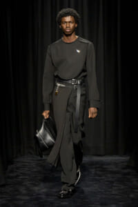 Male model in an all-black ensemble walks a runway, carrying a black leather bag and wearing a wide belt with metal hardware.