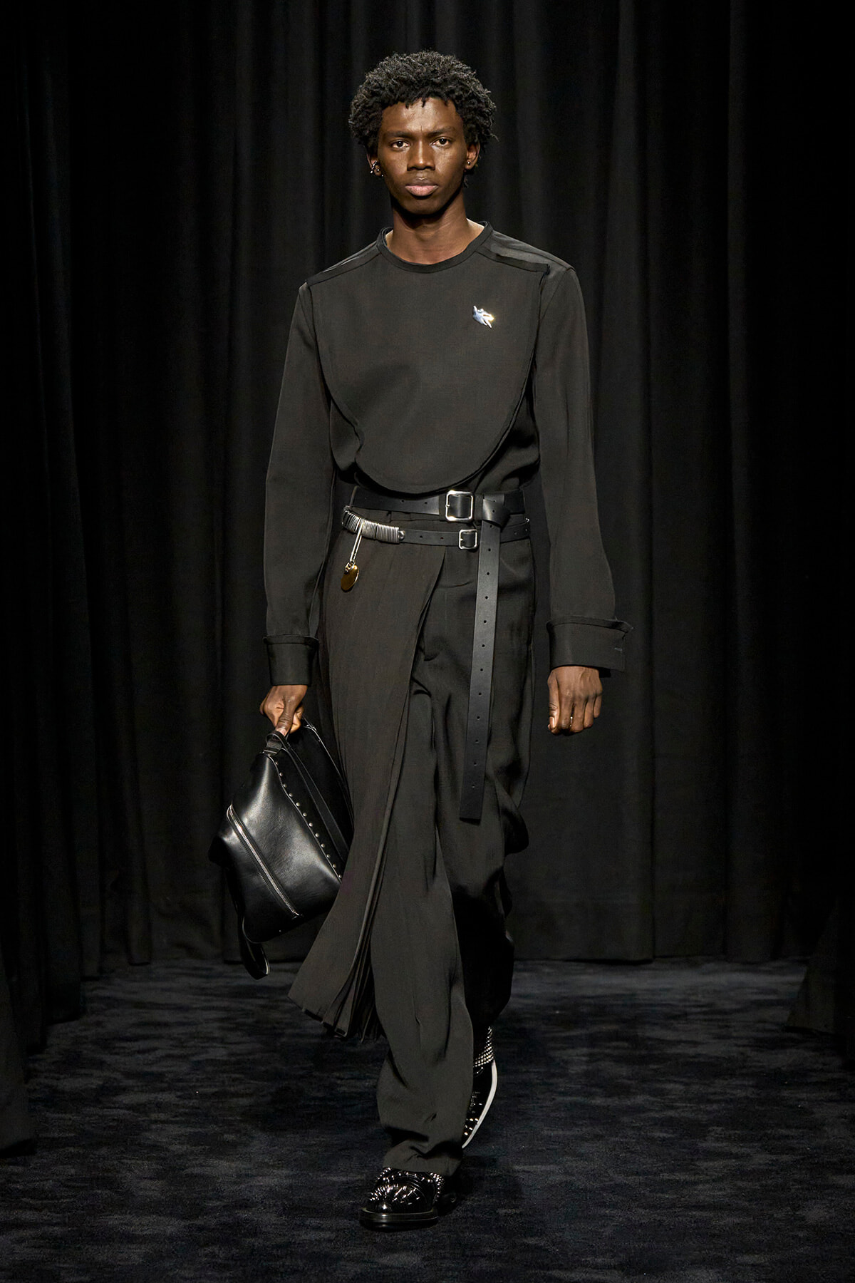 Male model in an all-black ensemble walks a runway, carrying a black leather bag and wearing a wide belt with metal hardware.