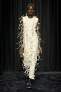 Model on a dark runway wearing a white sleeveless dress with cascading fringe strands all over, a beige clutch in hand, and black studded shoes.