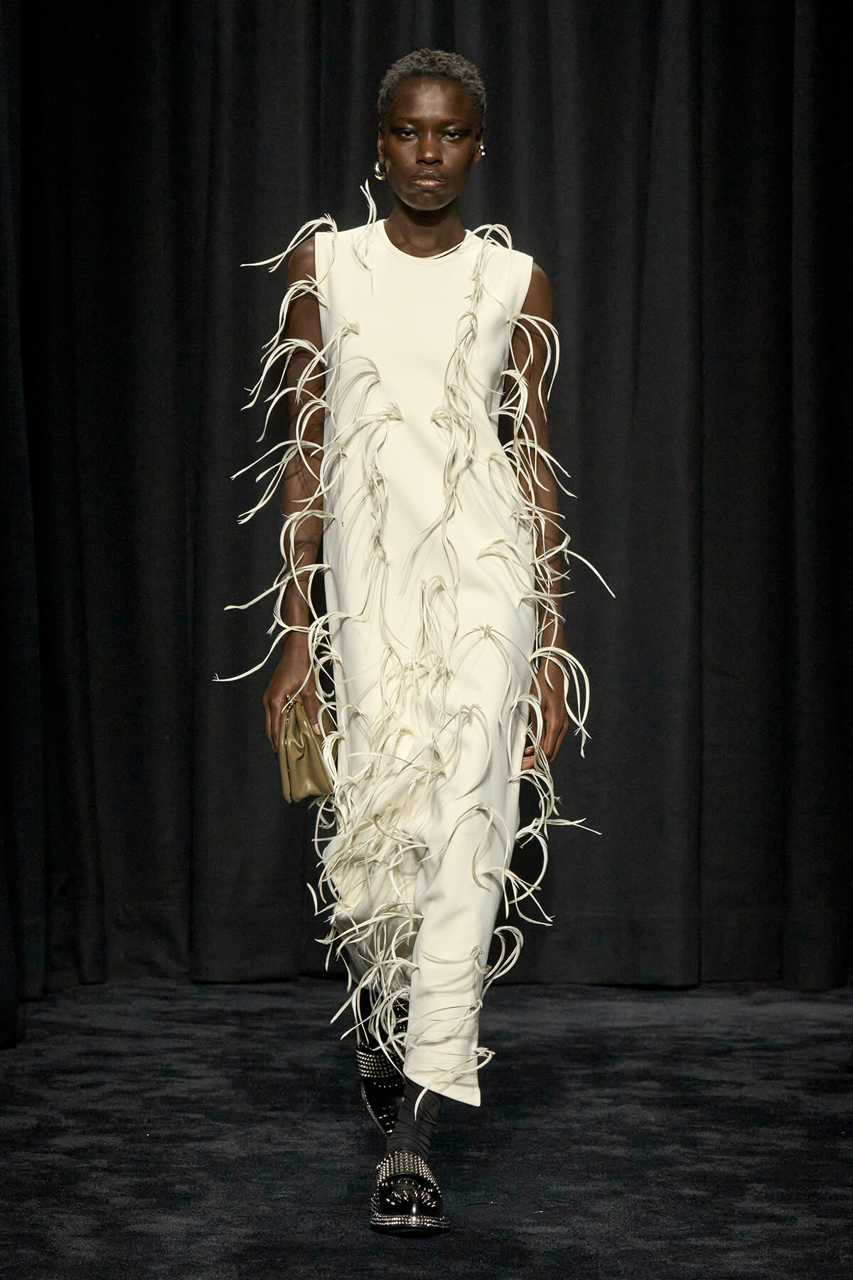 Model on a dark runway wearing a white sleeveless dress with cascading fringe strands all over, a beige clutch in hand, and black studded shoes.
