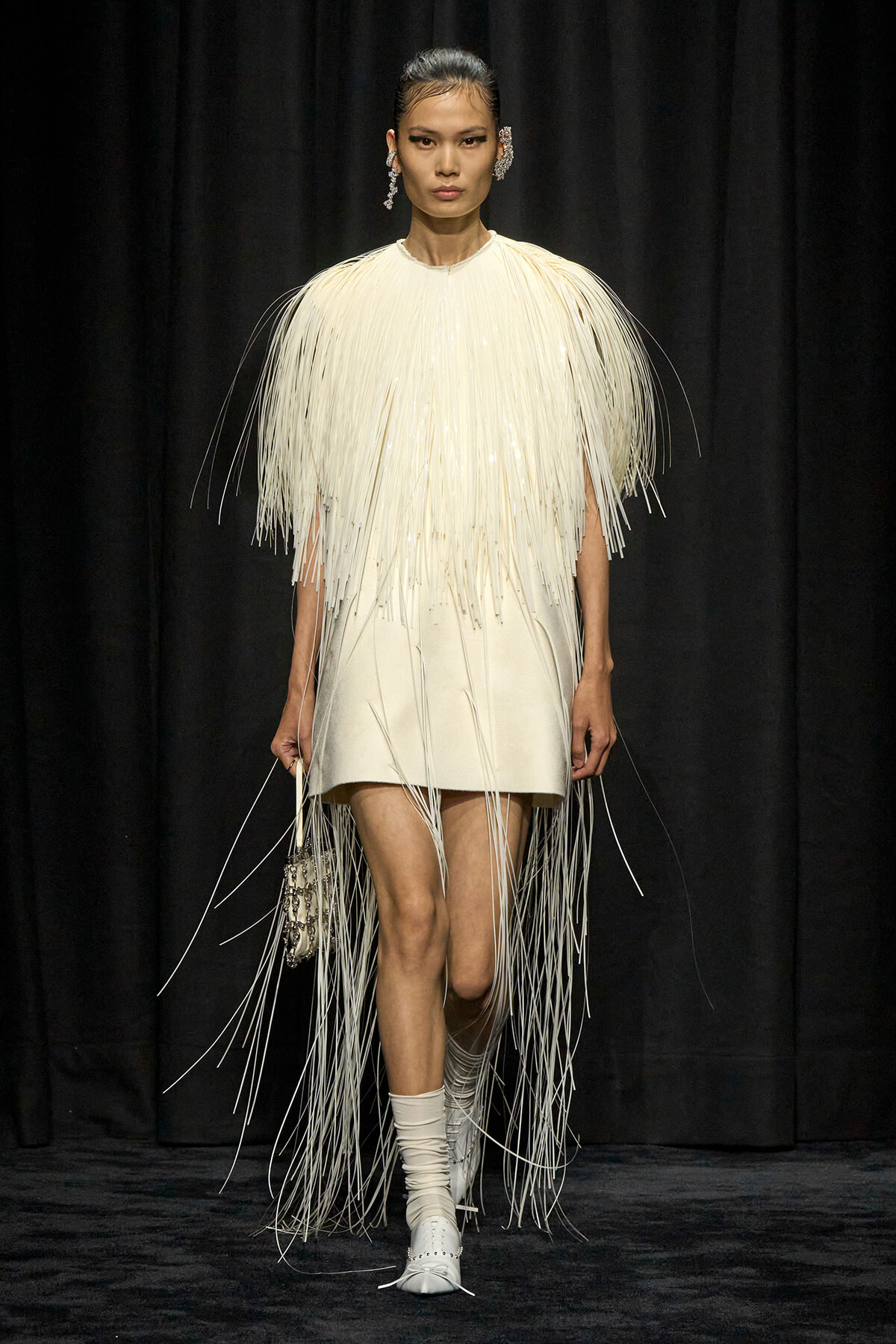 Model walking a runway in a cream fringe dress with long strand embellishments and matching accessories.