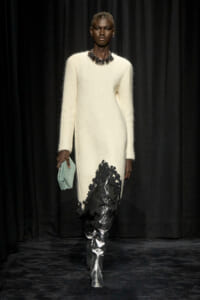 Model walking a runway in a cream knit dress with a black floral-appliqué jet-black side panel, accessorized with a statement necklace and mint clutch.