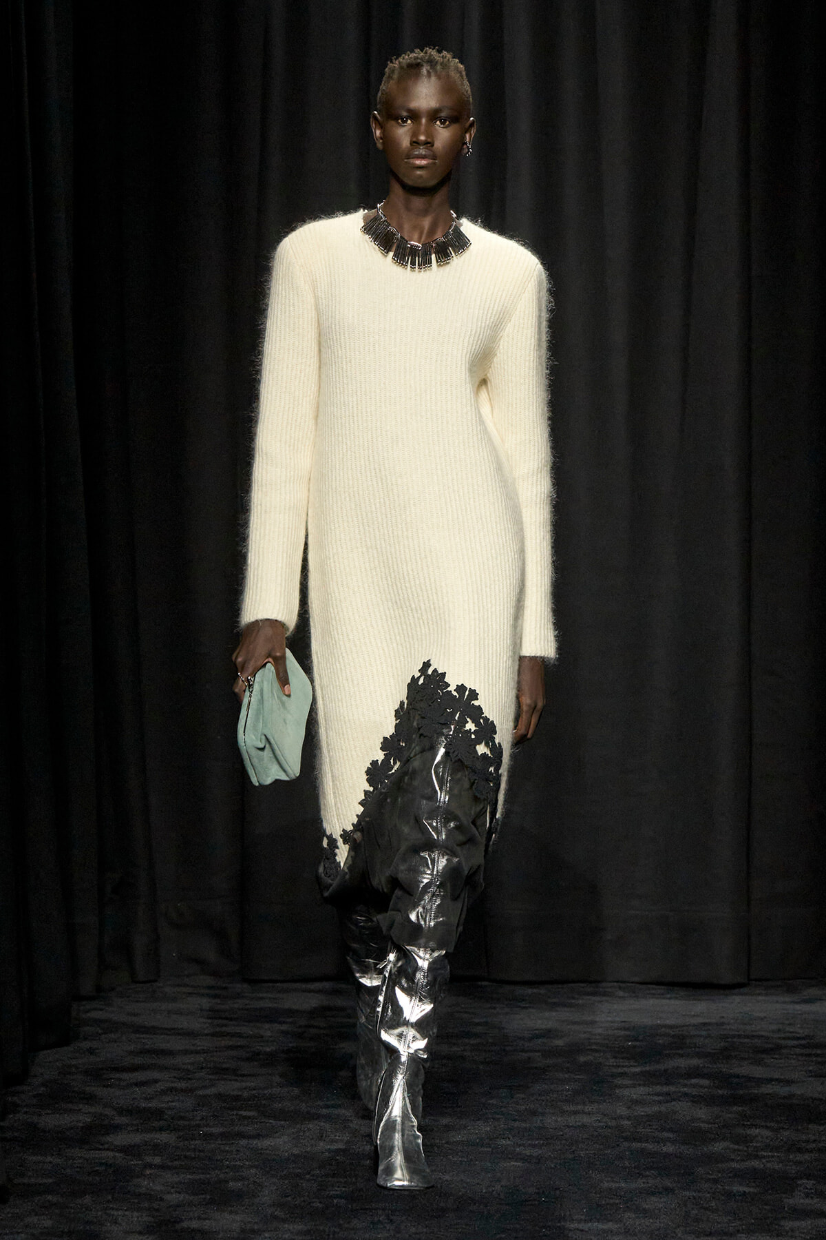 Model walking a runway in a cream knit dress with a black floral-appliqué jet-black side panel, accessorized with a statement necklace and mint clutch.