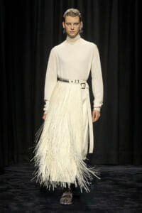 Runway model in an ivory turtleneck sweater with a long, fringed cream skirt and a metallic belt, standing against a black backdrop with bold earrings in place.