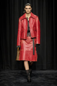 Model walks the runway in a red leather suit: oversized jacket, matching knee-length skirt, black top, wide belt, and brown handbag.
