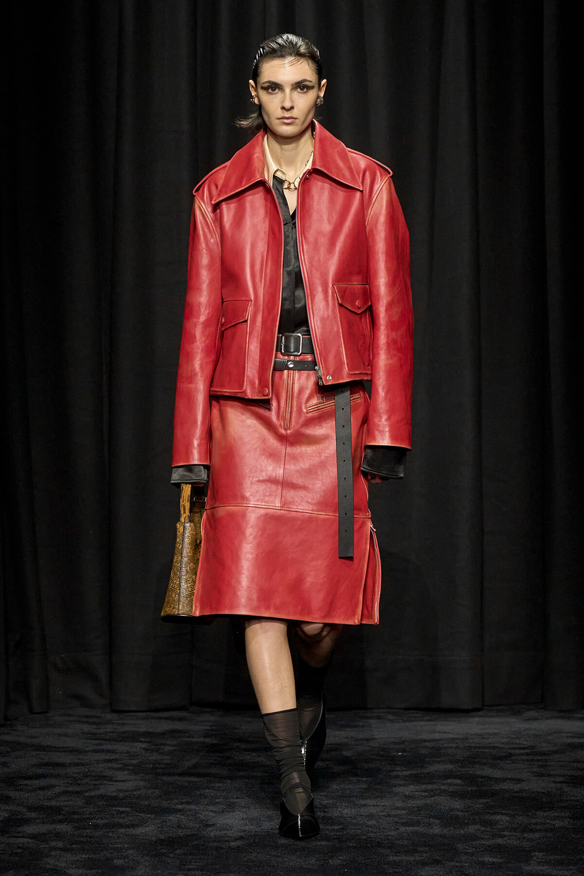 Model walks the runway in a red leather suit: oversized jacket, matching knee-length skirt, black top, wide belt, and brown handbag.