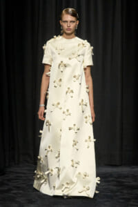 Model on a runway wearing a cream ankle-length dress decorated with scattered ribbon bows against a dark backdrop.