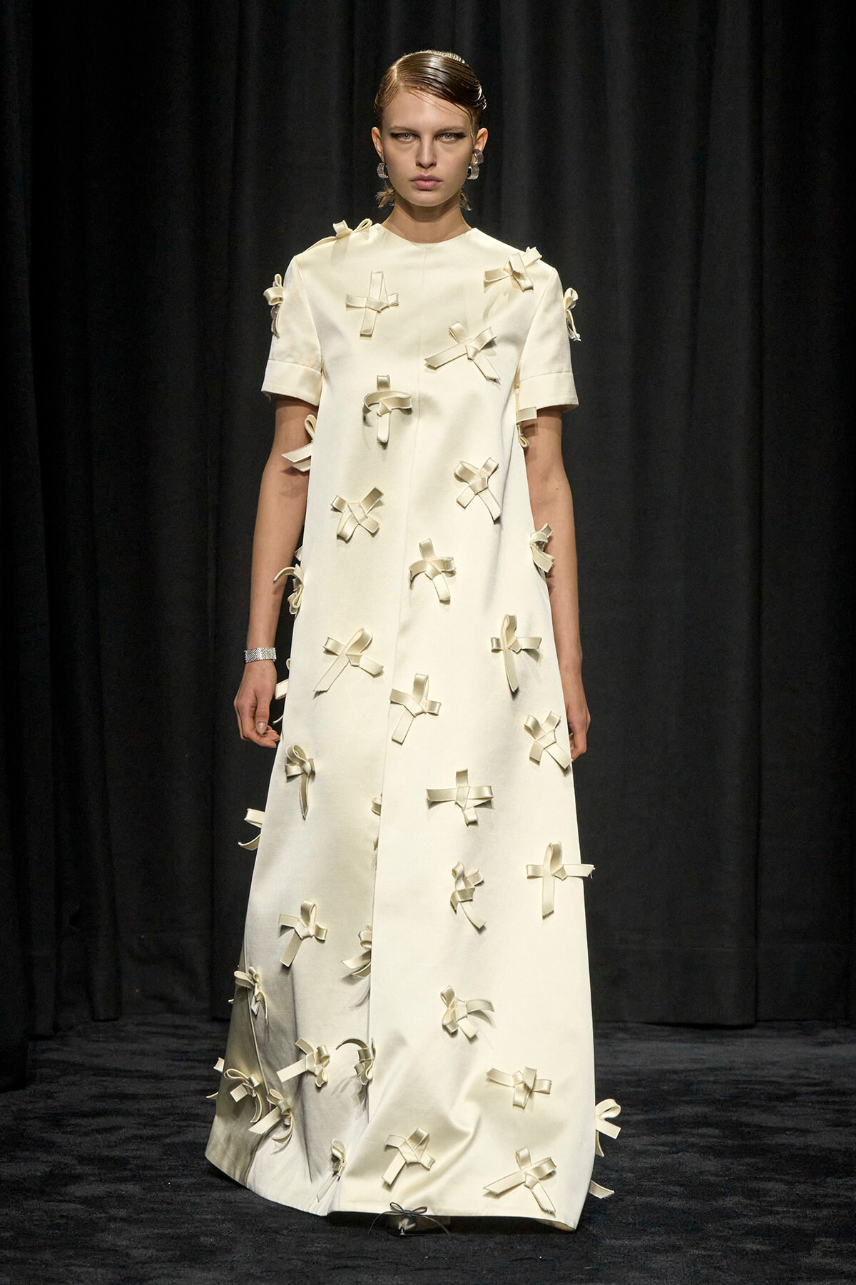 Model on a runway wearing a cream ankle-length dress decorated with scattered ribbon bows against a dark backdrop.