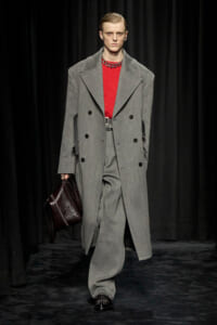Model on a runway wearing a gray oversized double-breasted coat over a red knit sweater, with a dark leather briefcase.