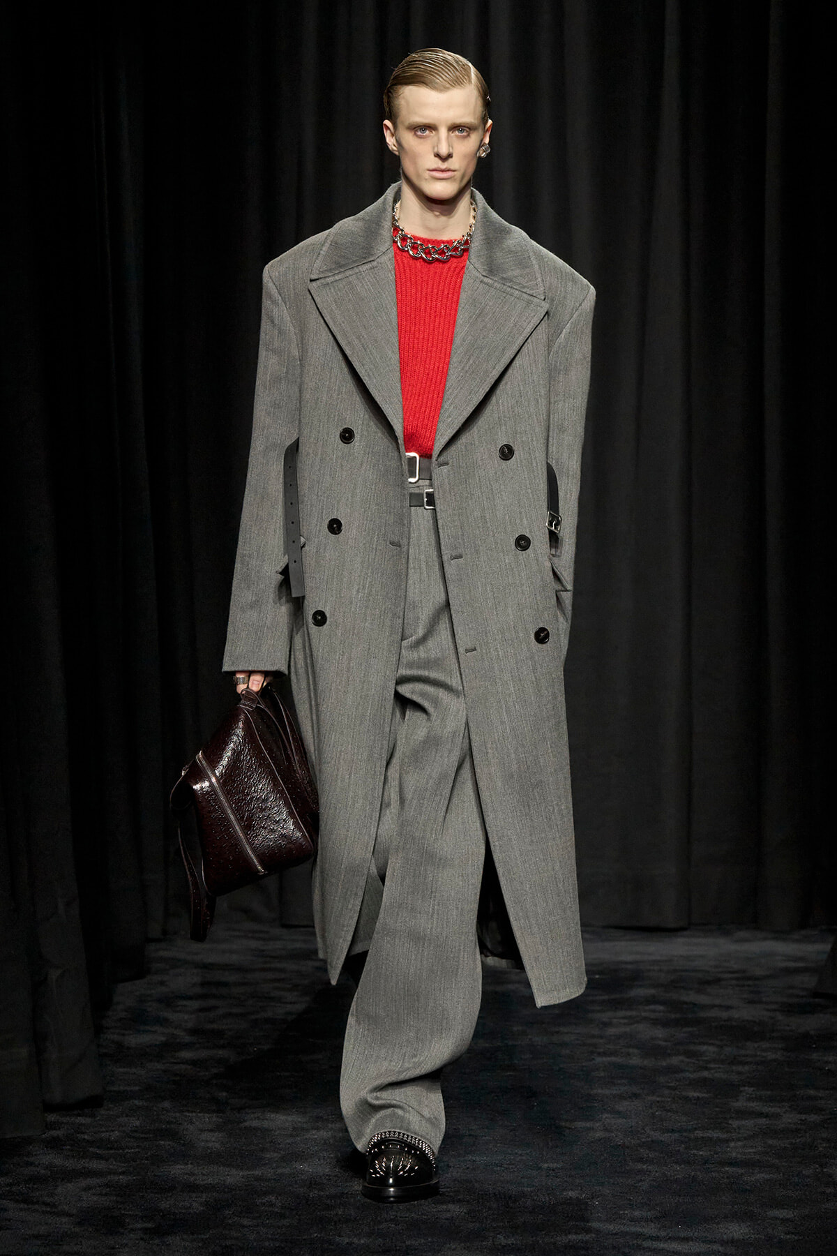 Model on a runway wearing a gray oversized double-breasted coat over a red knit sweater, with a dark leather briefcase.