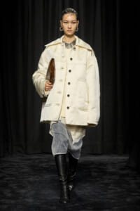 Runway model in an oversized cream coat with large pockets, holding a brown clutch, paired with draped gray pants and black knee-high boots.