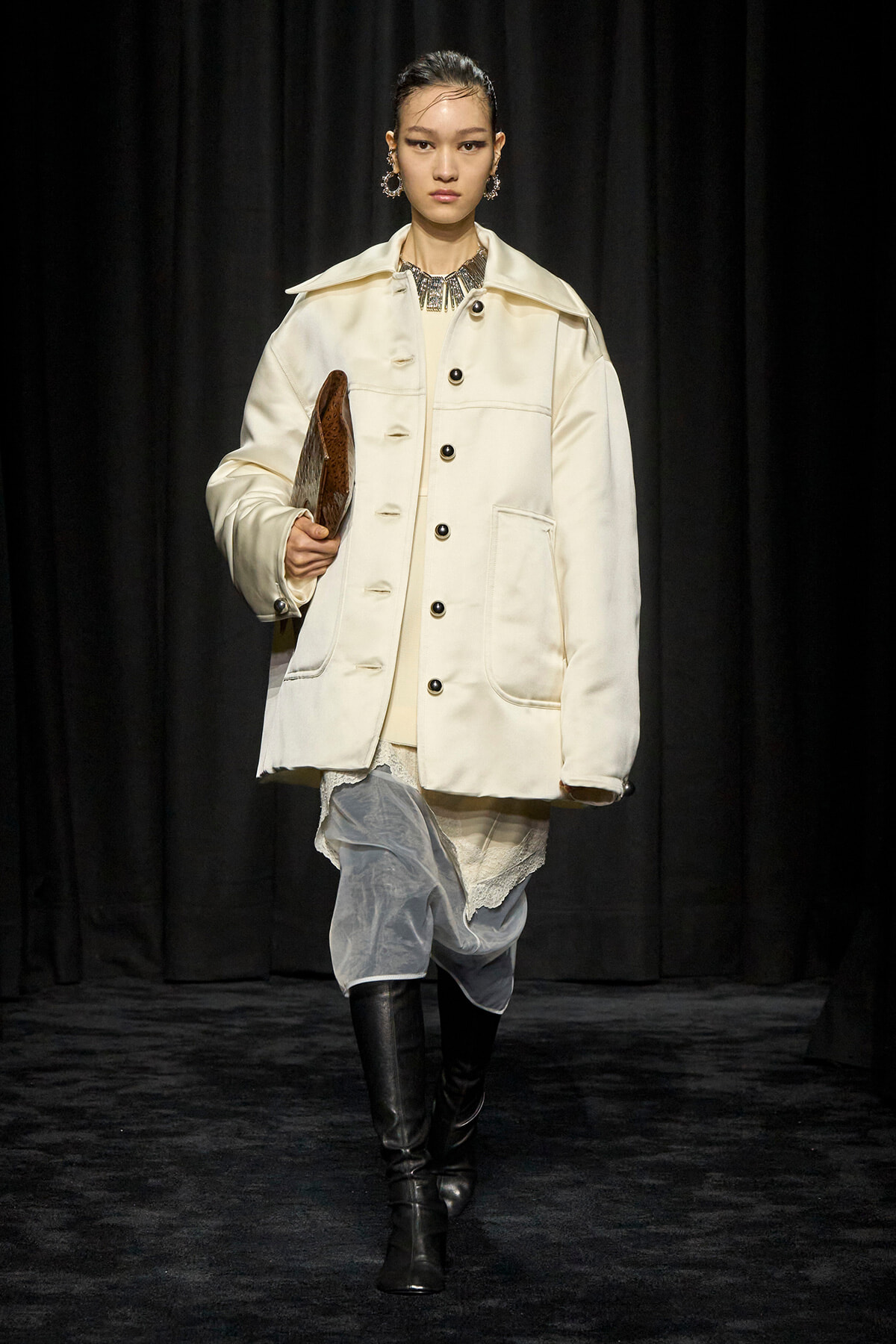 Runway model in an oversized cream coat with large pockets, holding a brown clutch, paired with draped gray pants and black knee-high boots.