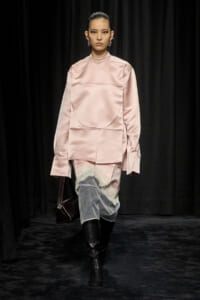 Model on a runway wearing a pale pink satin oversized top with a curved panel, paired with a gray lace-trimmed skirt and black knee-high boots.