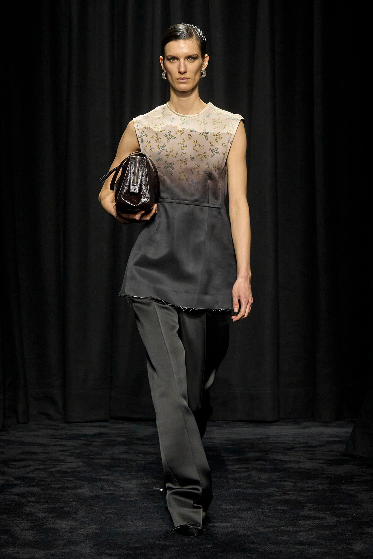 Model walks the runway in a sleeveless two-tone top with delicate floral embroidery, paired with gray wide-leg pants, carrying a small dark handbag.