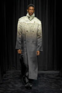 Model on a runway wearing a knee-length gradient trench coat fading from cream to dark gray, with a subtle floral pattern and high collar.