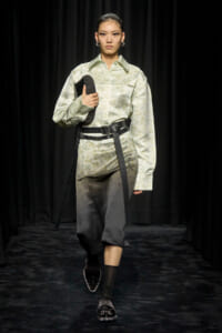 Model on a runway wearing a pale green floral shirt with a wide black belt, carrying a black studded clutch.