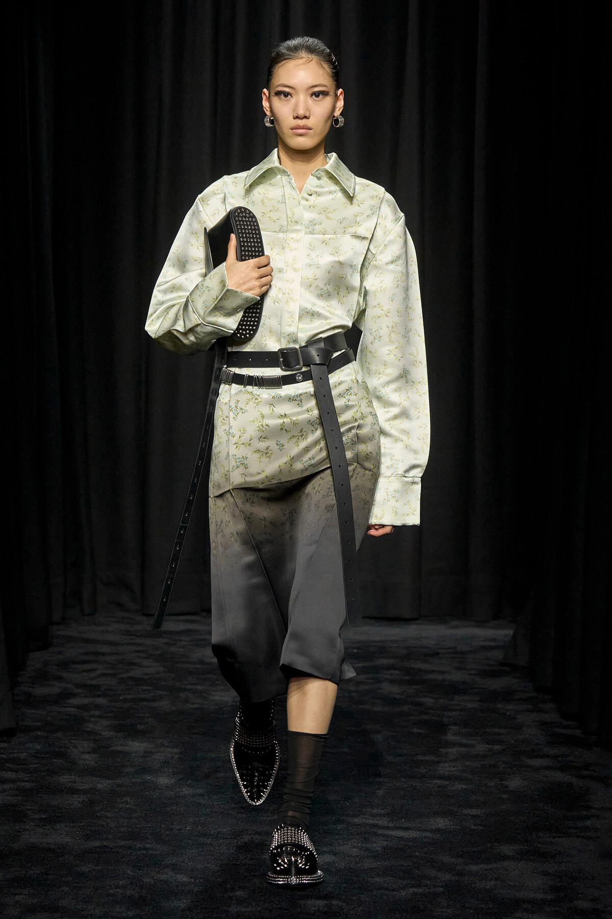 Model on a runway wearing a pale green floral shirt with a wide black belt, carrying a black studded clutch.