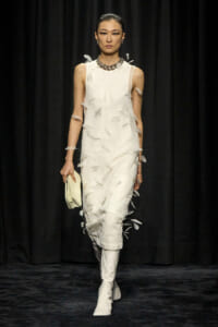 Runway model in an ivory sleeveless dress with feather appliqués, paired with white knee-high boots and a chunky necklace, holding a pale clutch.