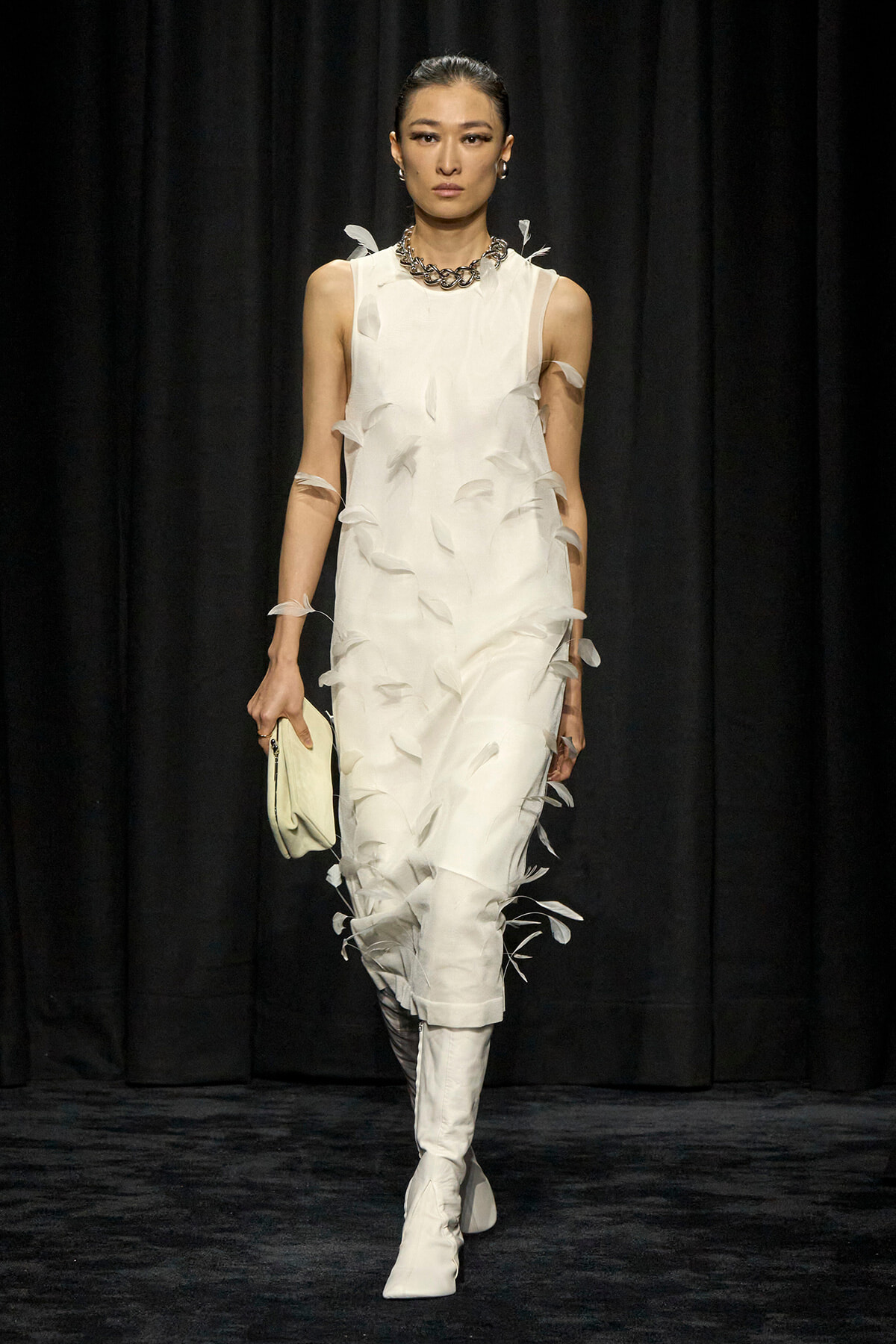 Runway model in an ivory sleeveless dress with feather appliqués, paired with white knee-high boots and a chunky necklace, holding a pale clutch.