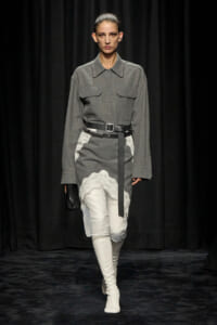 Runway model in a gray belted shirt dress with lace hem over white pants and white knee-high boots, holding a black clutch.