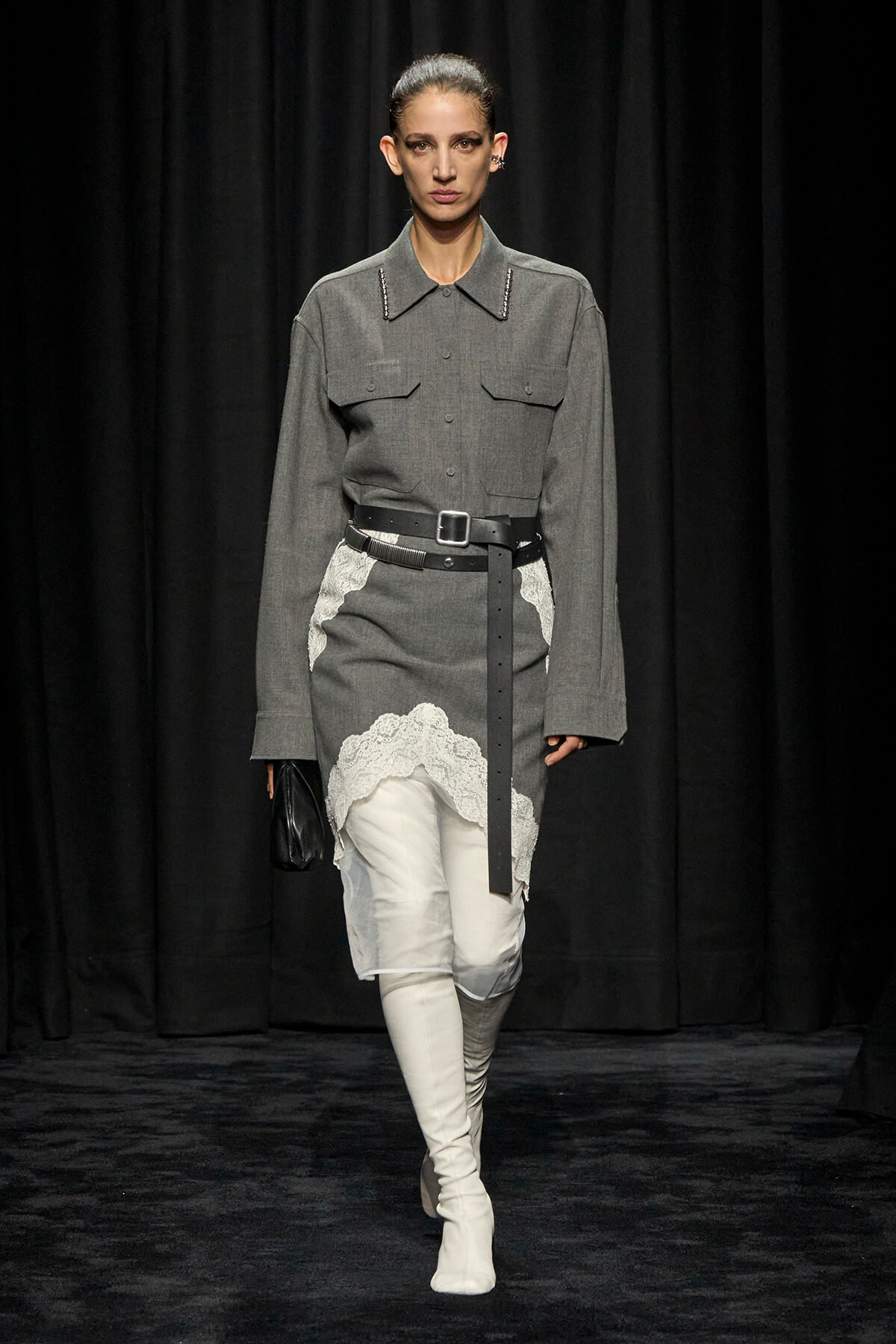 Runway model in a gray belted shirt dress with lace hem over white pants and white knee-high boots, holding a black clutch.