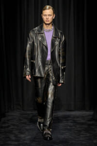 Model walking a runway in a black leather biker jacket and matching pants with a purple knit top and metallic shoes on a dark backdrop by curtains.
