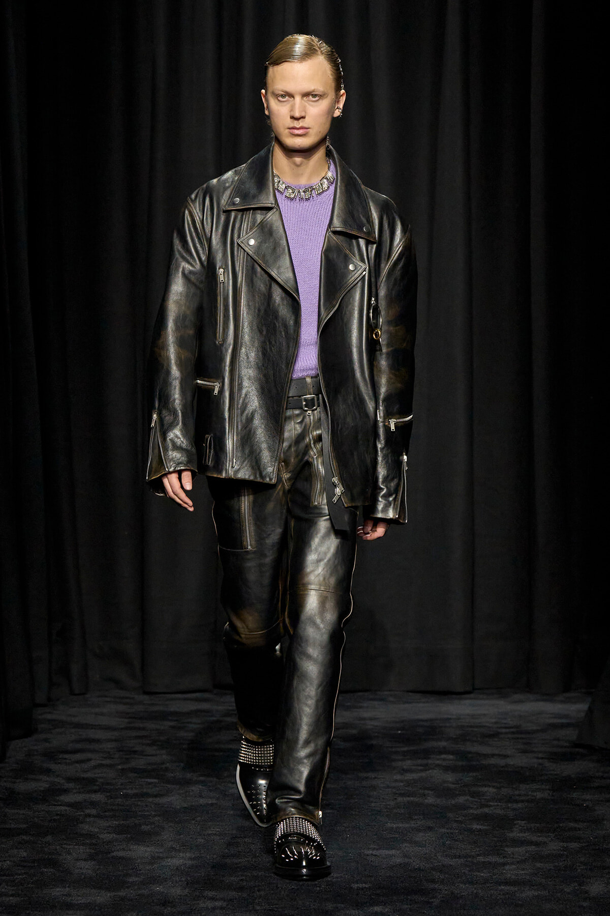 Model walking a runway in a black leather biker jacket and matching pants with a purple knit top and metallic shoes on a dark backdrop by curtains.