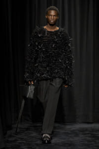 Model on a dark runway wearing an oversized black textured knit top with fringe, wide-leg black trousers, studded black loafers, and a small structured black bag.