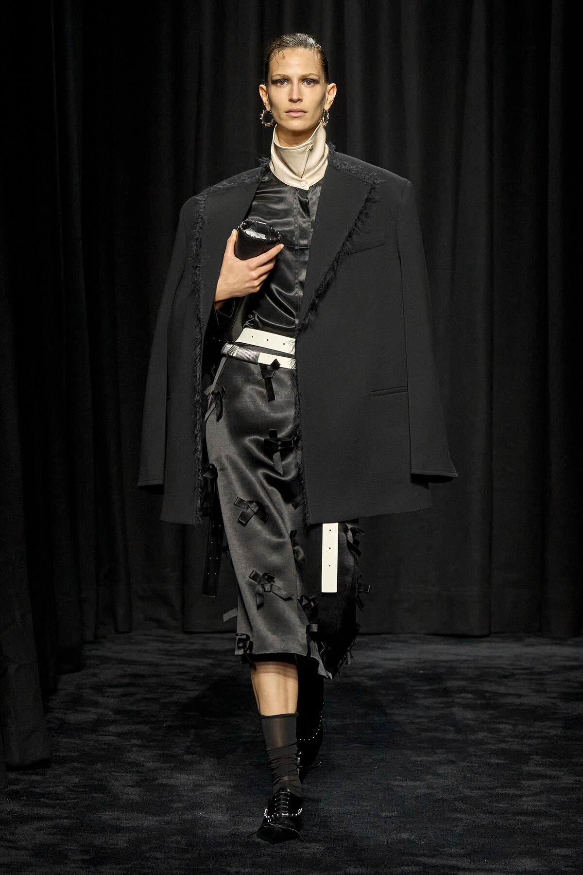 Model on a dim runway wearing a black oversized blazer over a dark satin dress with decorative bows, white belt, and black ankle boots, clutch in hand.