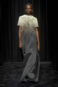 Model walking a fashion show in a two-tone gown: pale embroidered bodice that fades into a dark grey satin skirt, with short sleeves and silver jewelry against a black backdrop.