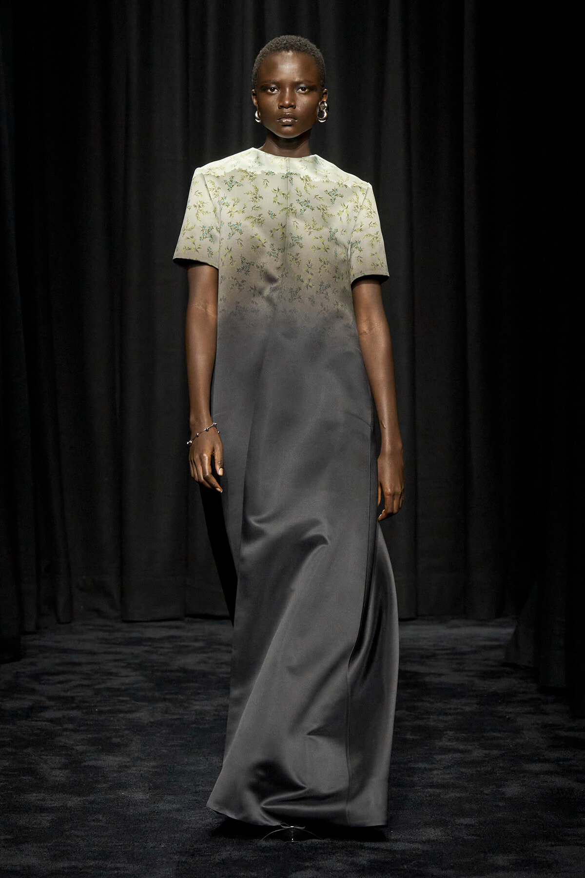 Model walking a fashion show in a two-tone gown: pale embroidered bodice that fades into a dark grey satin skirt, with short sleeves and silver jewelry against a black backdrop.
