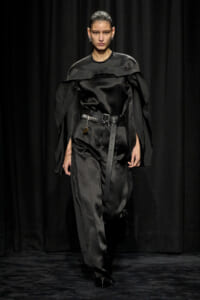 Model walks a runway in a black satin jumpsuit with cape-like shoulders and a wide belt, against a dark backdrop.
