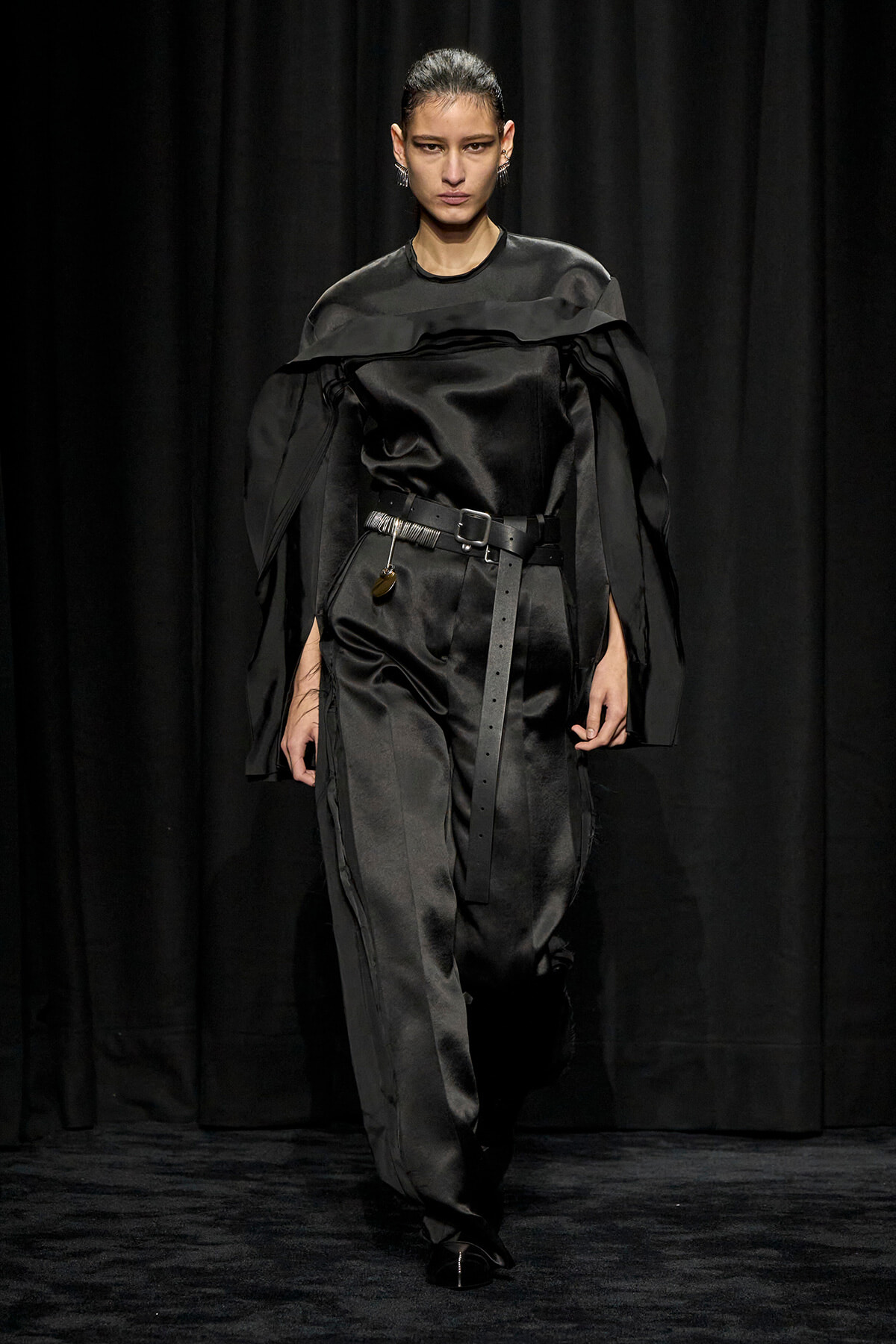 Model walks a runway in a black satin jumpsuit with cape-like shoulders and a wide belt, against a dark backdrop.