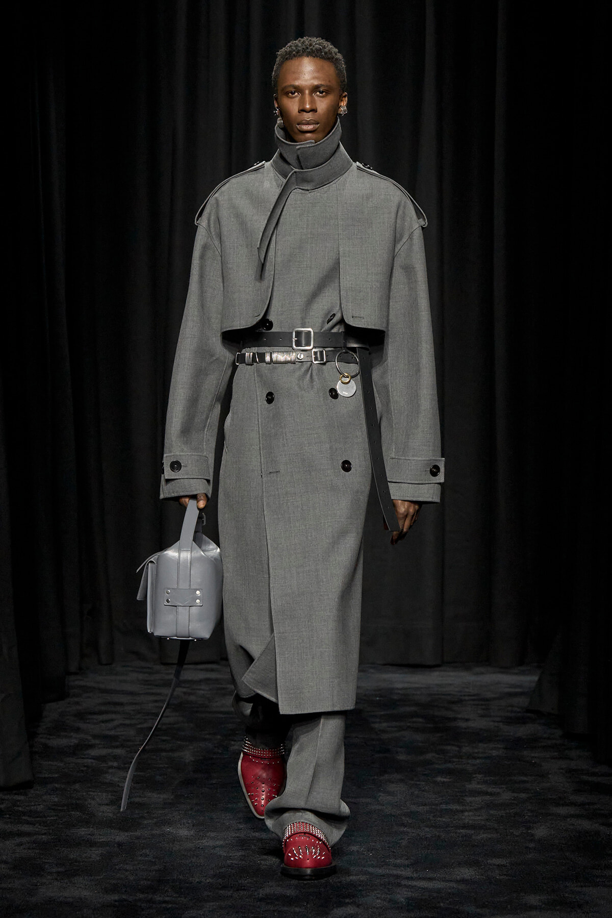 Male model walks a runway in a long grey trench coat with a waist belt, paired with red studded shoes and a small grey bag.