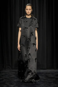 Model on a runway wearing a dark gray, floor-length dress adorned with numerous black ribbon bows on a black backdrop.