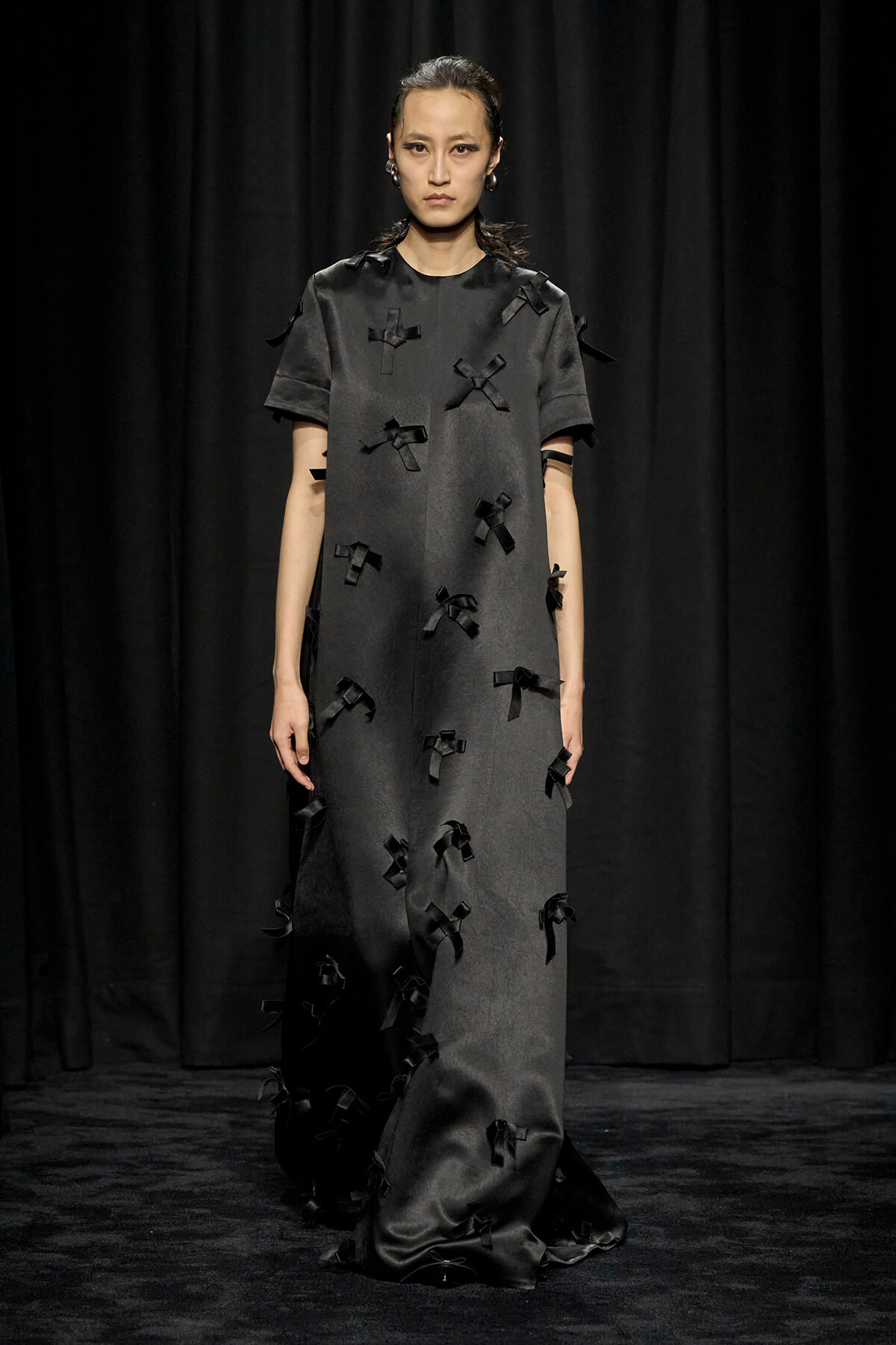Model on a runway wearing a dark gray, floor-length dress adorned with numerous black ribbon bows on a black backdrop.