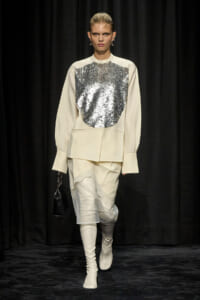 Model on a runway wearing an ivory oversized top with a large silver sequin panel, a cream skirt with lace details, white knee-high boots, and a small black handbag.