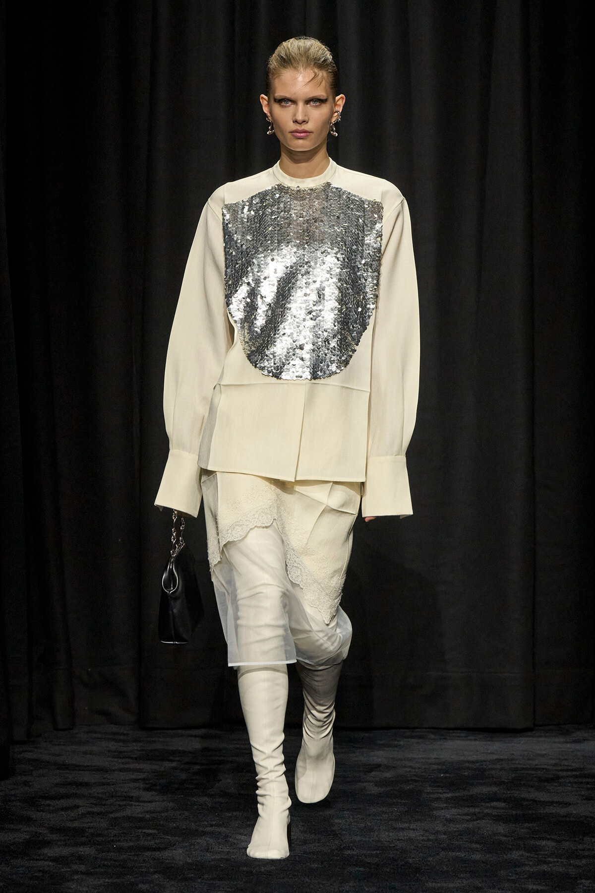 Model on a runway wearing an ivory oversized top with a large silver sequin panel, a cream skirt with lace details, white knee-high boots, and a small black handbag.