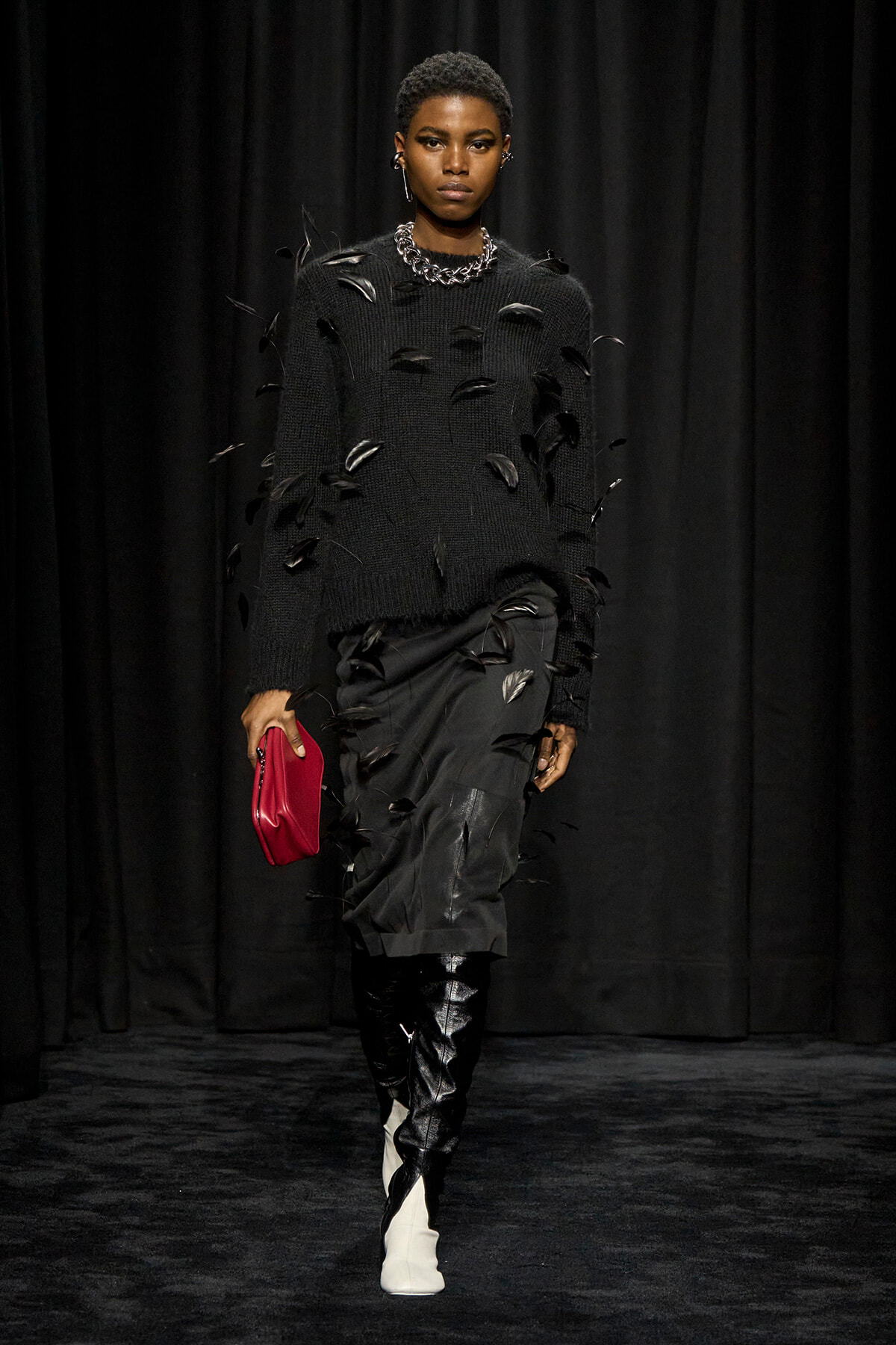 Model on a dark runway wearing a black knit sweater with feather embellishments, black skirt, white knee boots, a chunky silver necklace, and a pink clutch.