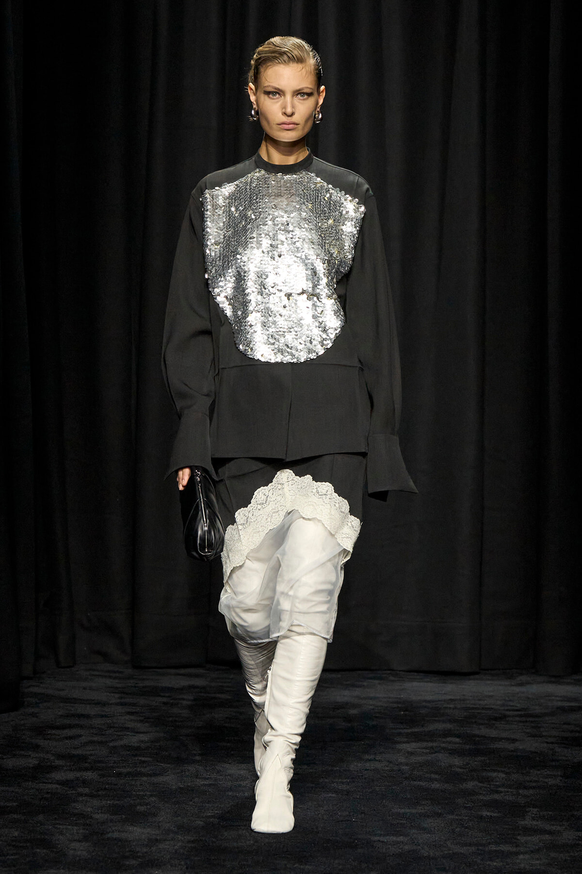 Model walking the runway in a black outfit with a large silver sequin front panel, white lace-trimmed shorts, and white knee-high boots, carrying a small black clutch