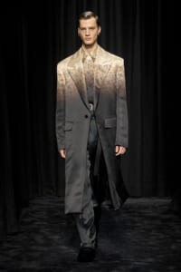 Male model walking a runway in a two-tone gradient suit jacket fading from beige floral pattern to dark gray, matching pants, black shoes.