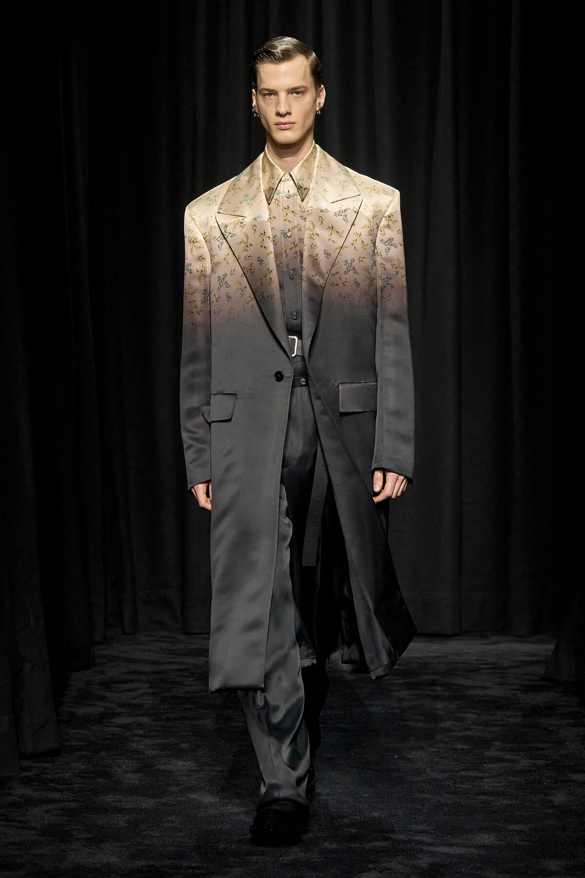 Male model walking a runway in a two-tone gradient suit jacket fading from beige floral pattern to dark gray, matching pants, black shoes.