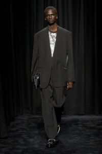 Male model on a runway wearing an oversized black suit with a silver sequined top, carrying a black clutch.