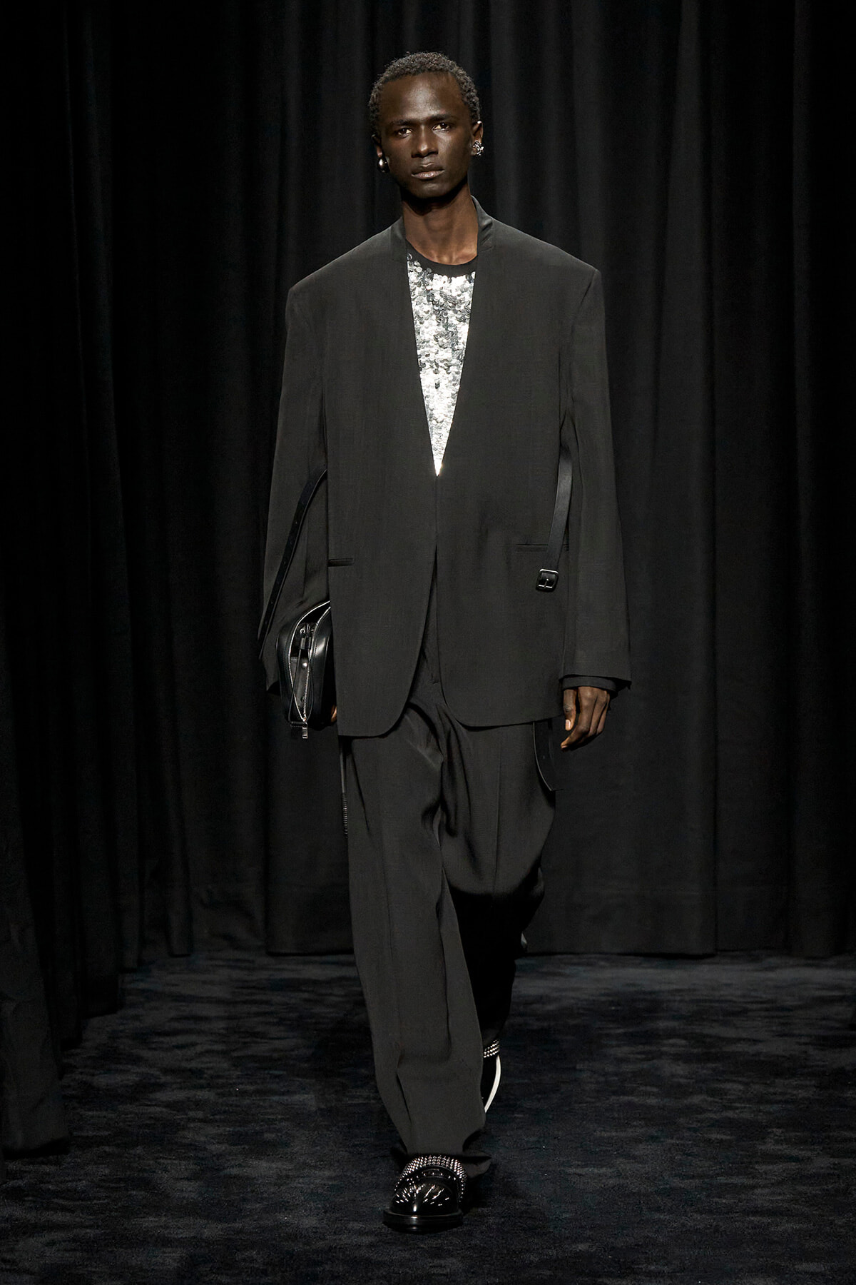 Male model on a runway wearing an oversized black suit with a silver sequined top, carrying a black clutch.