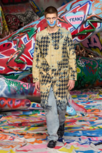 Model wearing oversized beige houndstooth coat with fringe, being blue-face painted on a graffiti-covered runway backdrop