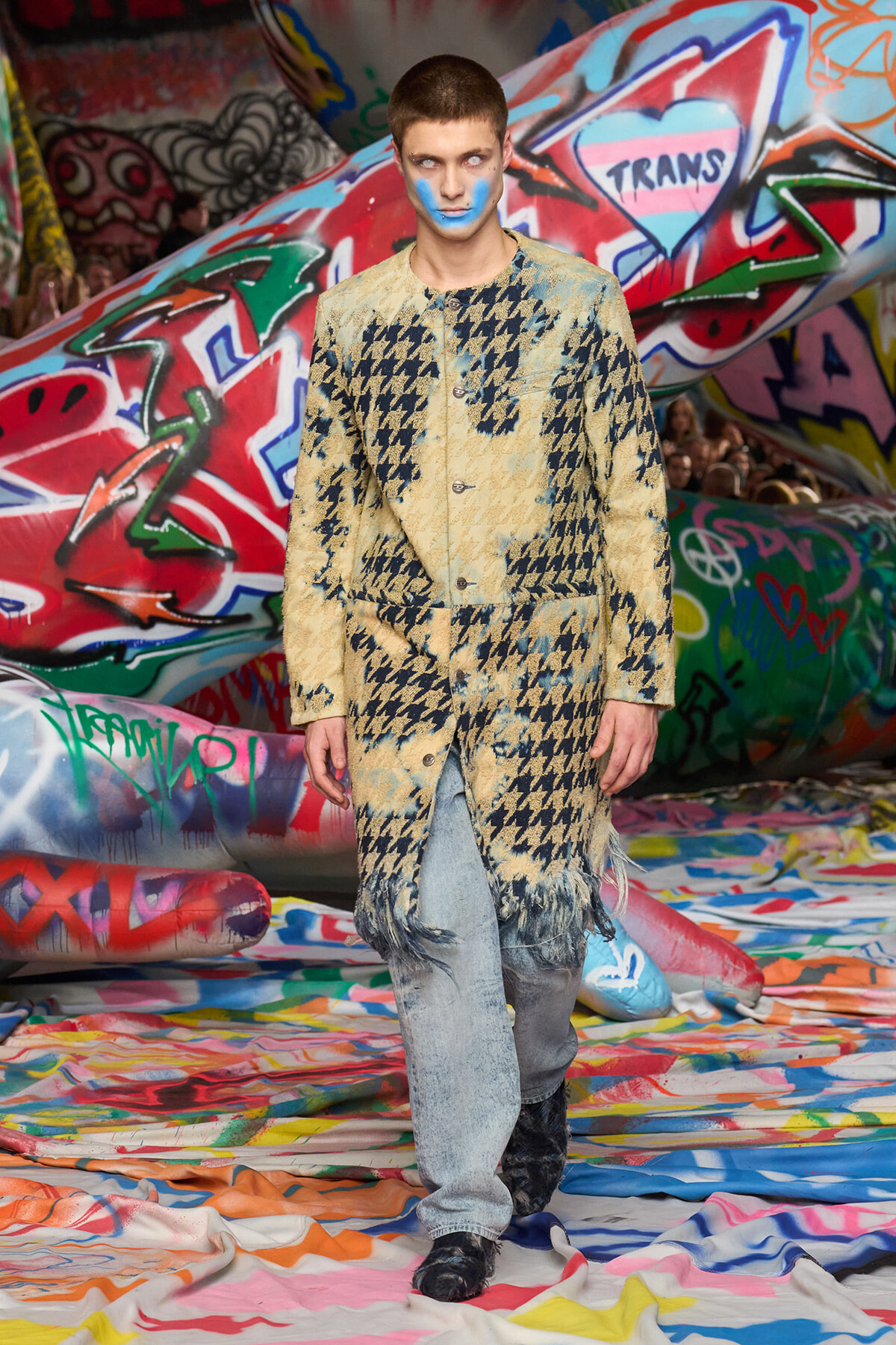 Model wearing oversized beige houndstooth coat with fringe, being blue-face painted on a graffiti-covered runway backdrop