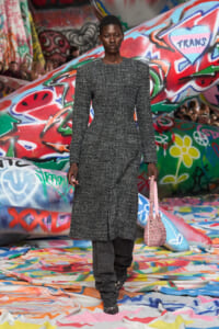 Model walking a runway in a gray textured coat and dark pants, holding a pink woven handbag against a colorful graffiti backdrop.