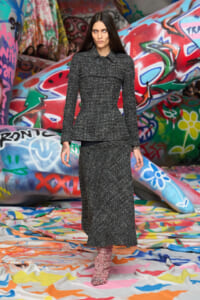 Model walking the runway in a dark gray speckled tweed suit with a fitted blazer and ankle-length skirt, set against colorful graffiti walls.