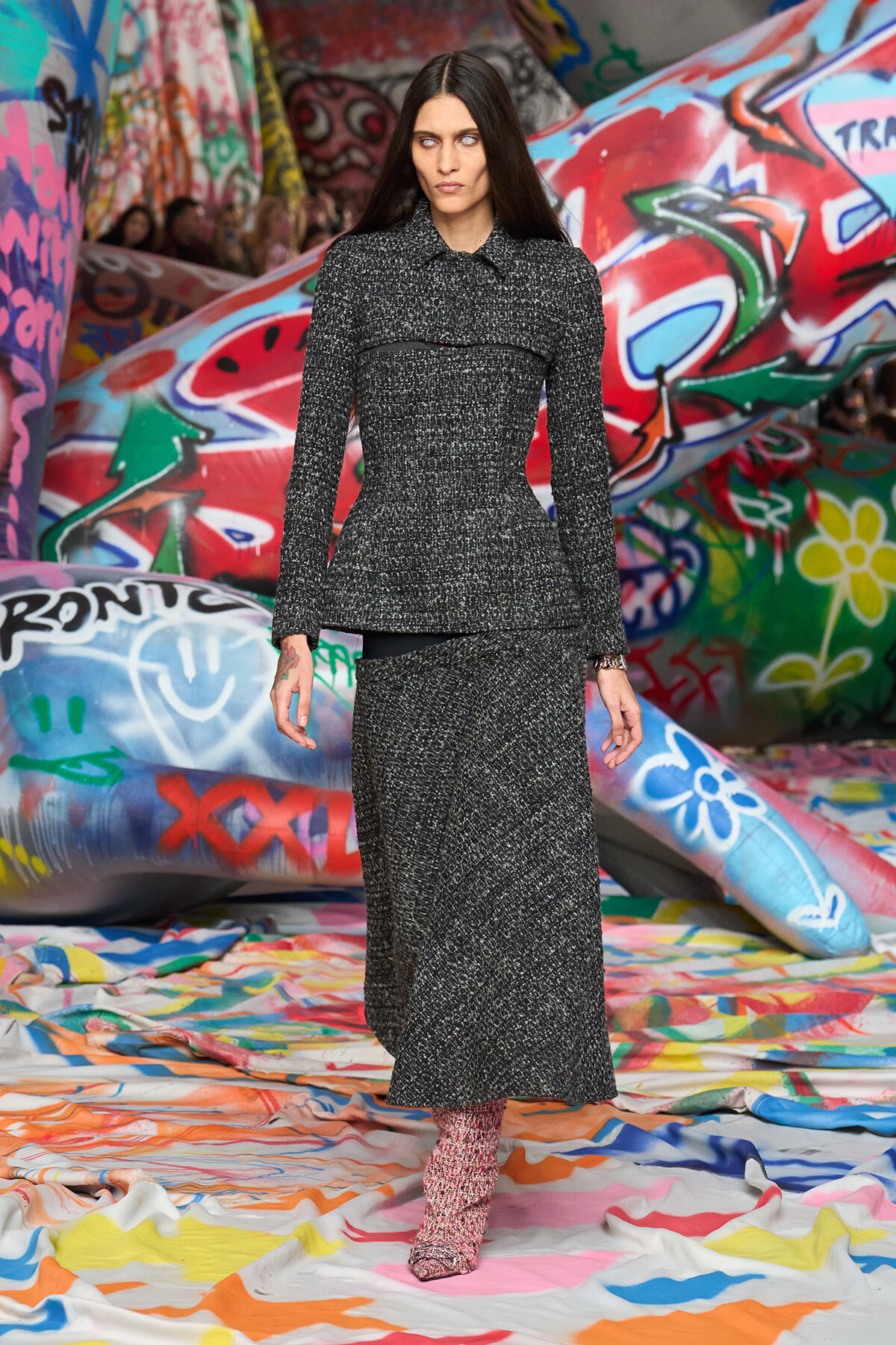 Model walking the runway in a dark gray speckled tweed suit with a fitted blazer and ankle-length skirt, set against colorful graffiti walls.